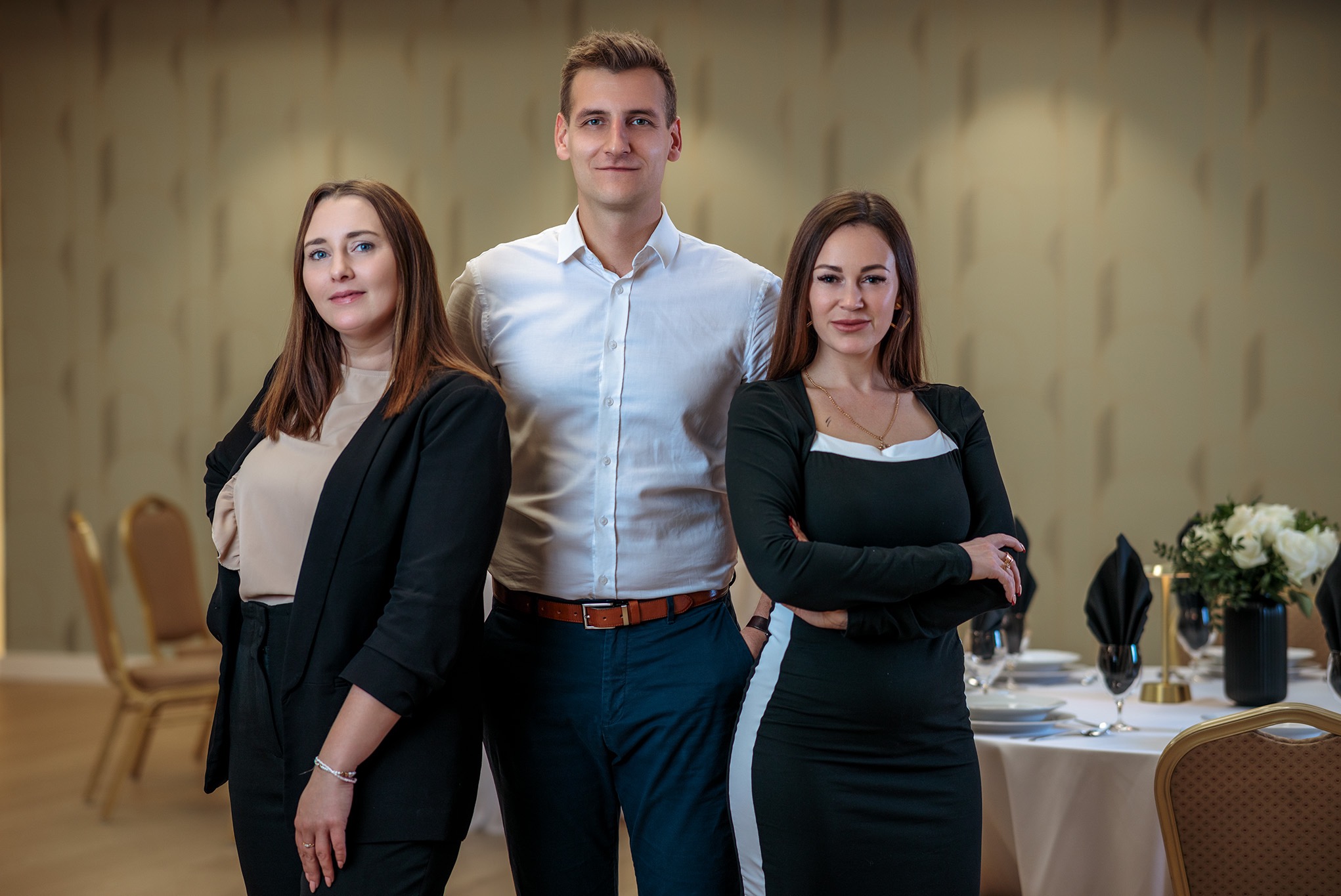 Portret grupowy: mężczyzna w białej koszuli i dwie kobiety w eleganckich strojach na tle sali bankietowej z nakrytymi stołami.