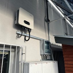 NASZA ENERGETYKA SPÓŁKA Z OGRANICZONĄ ODPOWIEDZIALNOŚCIĄ - Biały falownik fotowoltaiczny marki Sofar zamontowany na ścianie budynku, z widocznymi kablami, skrzynką elektryczną i panelami słonecznymi na dachu w tle.