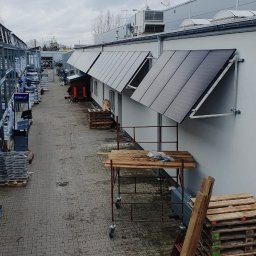 NASZA ENERGETYKA SPÓŁKA Z OGRANICZONĄ ODPOWIEDZIALNOŚCIĄ - Panele fotowoltaiczne zamontowane na elewacji budynku przemysłowego, widok z góry, w tle widoczne materiały budowlane i rusztowania.