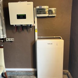 NASZA ENERGETYKA SPÓŁKA Z OGRANICZONĄ ODPOWIEDZIALNOŚCIĄ - Biały magazyn energii Sofar z podłączonym inwerterem i zabezpieczeniami, zamontowany na brązowej ścianie w piwnicy, widoczne okablowanie i elementy instalacji elektrycznej.