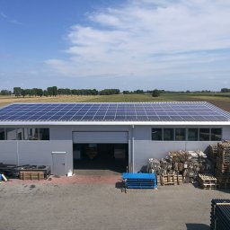NASZA ENERGETYKA SPÓŁKA Z OGRANICZONĄ ODPOWIEDZIALNOŚCIĄ - Hala magazynowa z panelami fotowoltaicznymi na dachu, widok z góry na zaplecze z paletami i materiałami budowlanymi, pola uprawne w tle.