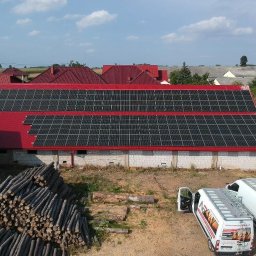 NASZA ENERGETYKA SPÓŁKA Z OGRANICZONĄ ODPOWIEDZIALNOŚCIĄ - Instalacja paneli fotowoltaicznych na czerwonym dachu budynku gospodarczego, widoczne dwa białe busy z otwartymi drzwiami, sterta pni drzew i osoba w tle.