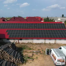 Instalacja paneli fotowoltaicznych na czerwonym dachu budynku gospodarczego, widoczne dwa białe busy z otwartymi drzwiami, sterta pni drzew i osoba w tle.