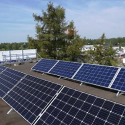 Panel słoneczny na dachu budynku, widok z góry, w tle drzewa i zabudowania miejskie.