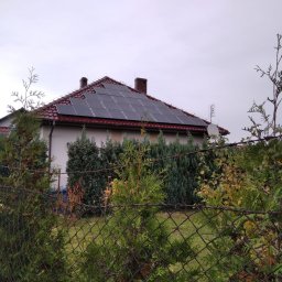 NASZA ENERGETYKA SPÓŁKA Z OGRANICZONĄ ODPOWIEDZIALNOŚCIĄ - Dom jednorodzinny z panelami fotowoltaicznymi na czerwonym dachu, widok przez zarośnięte ogrodzenie z siatki.