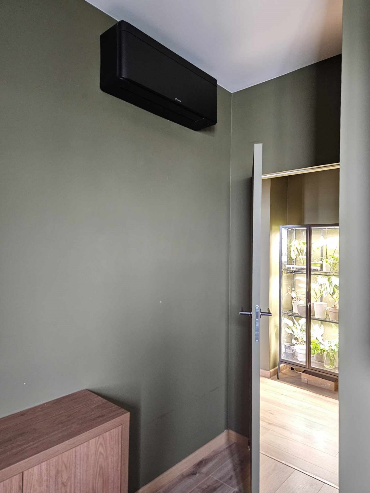Czarna jednostka klimatyzacyjna Daikin zamontowana na oliwkowozielonej ścianie, widok z dołu na otwarte drzwi z szafką na rośliny w tle.