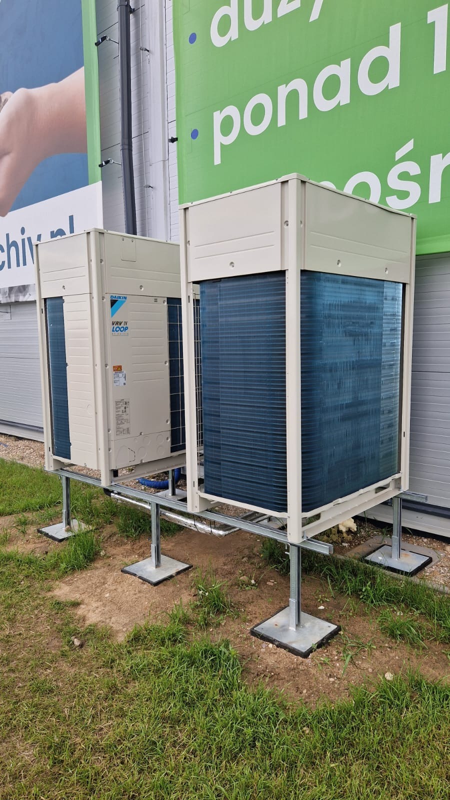 Dwie jednostki zewnętrzne klimatyzacji Daikin VRV IV LOOP zamontowane na metalowej konstrukcji wsporczej na trawniku przed budynkiem z reklamą.