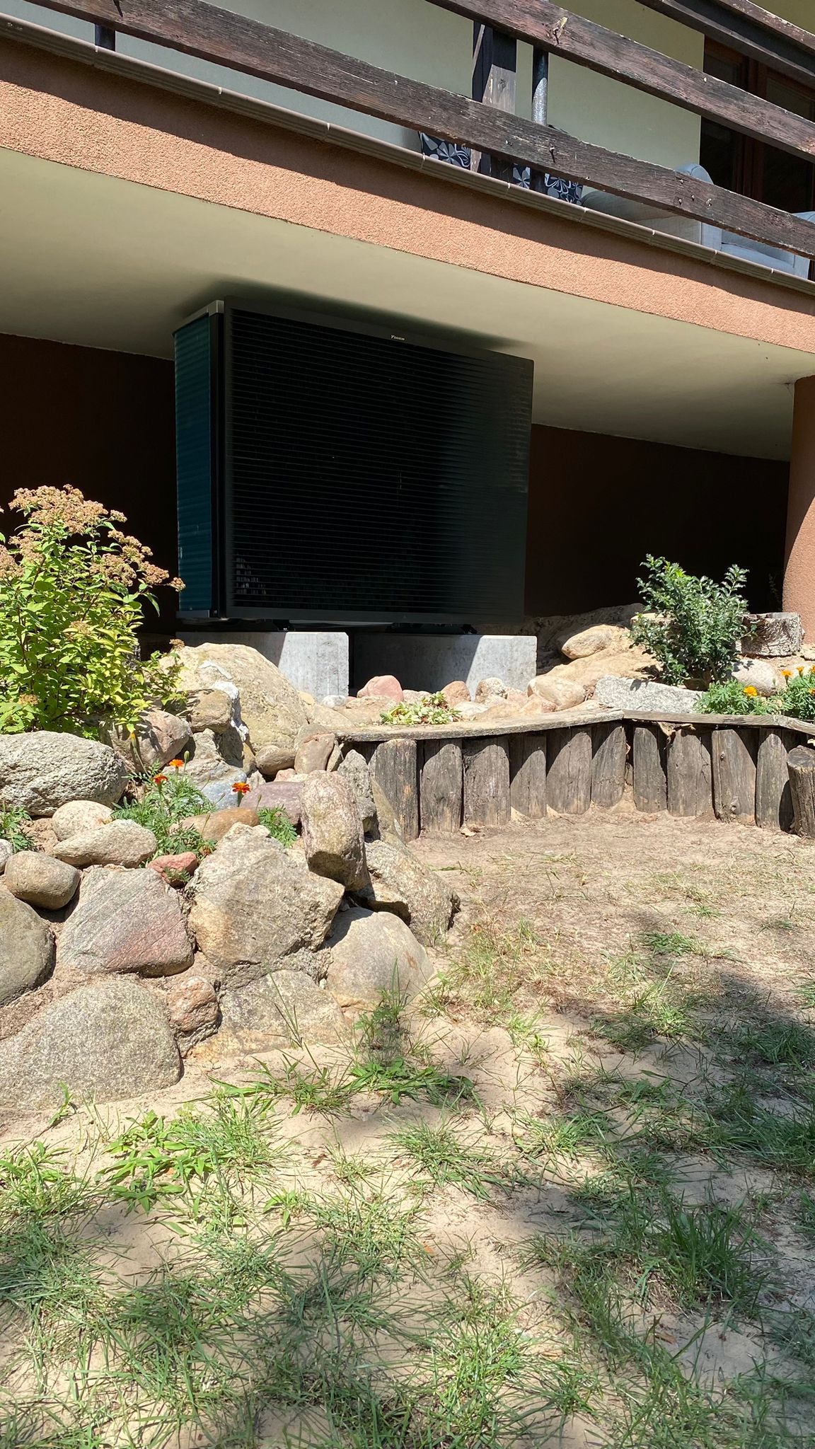 Zewnętrzna jednostka klimatyzacji Daikin zamontowana na betonowych blokach pod balkonem, otoczona skalniakiem z elementami drewnianymi.