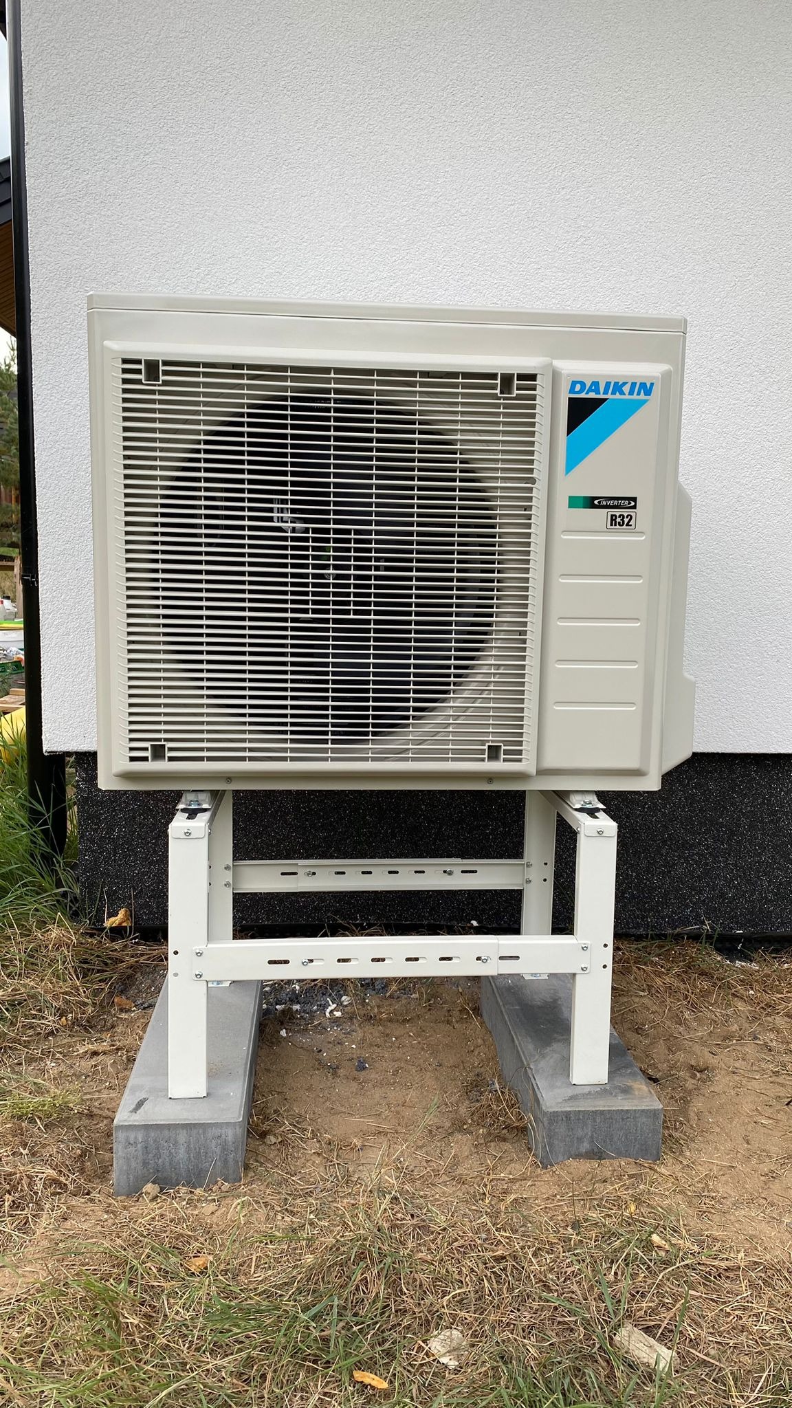 Zewnętrzna jednostka klimatyzacyjna Daikin R32 zamontowana na białym, metalowym stelażu na betonowych podkładach przy białej ścianie budynku.