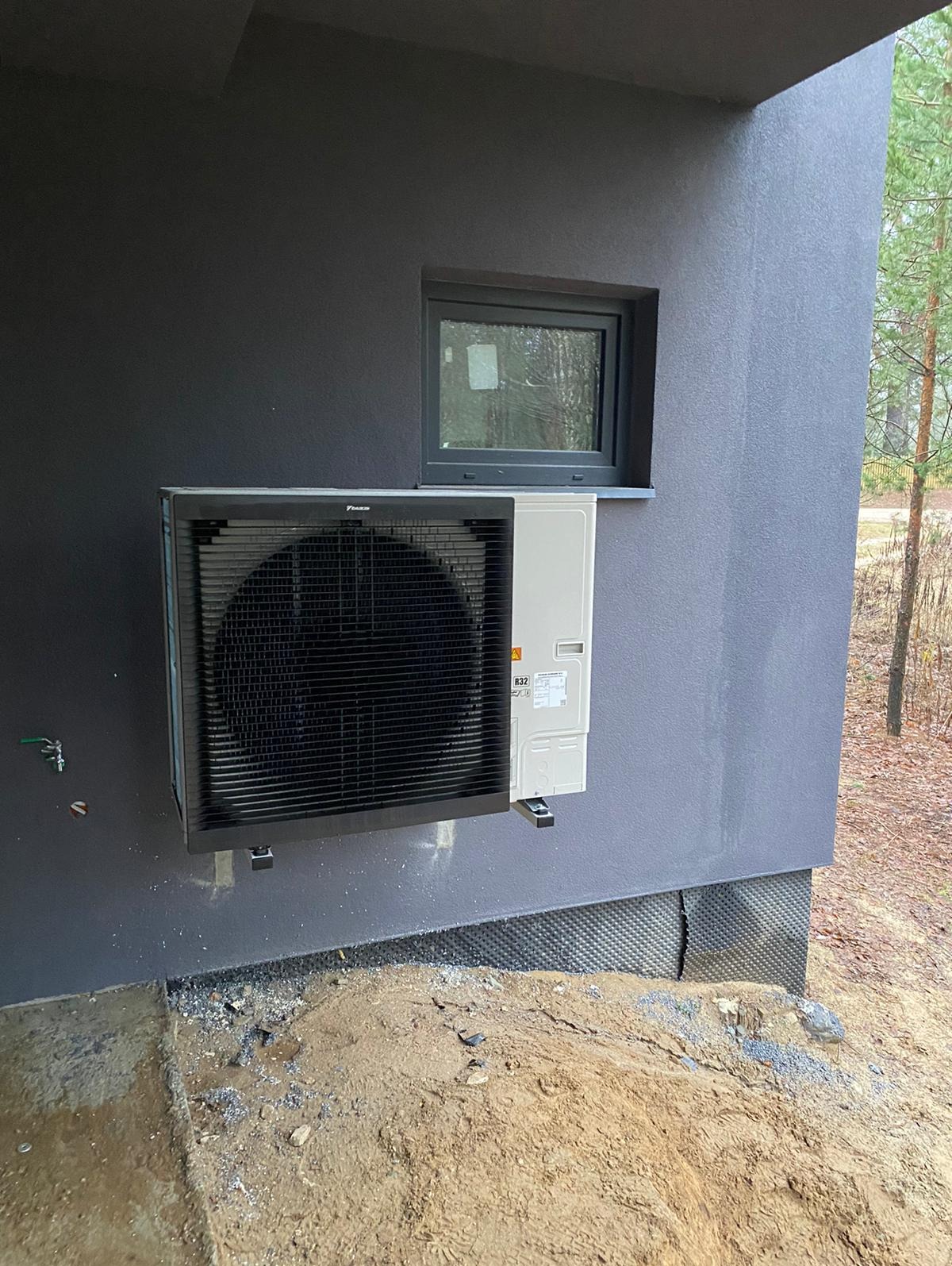 Zewnętrzna jednostka grzewcza Daikin zamontowana na szarej ścianie budynku obok okna, widoczny fragment terenu z piaskiem i elementami izolacji.