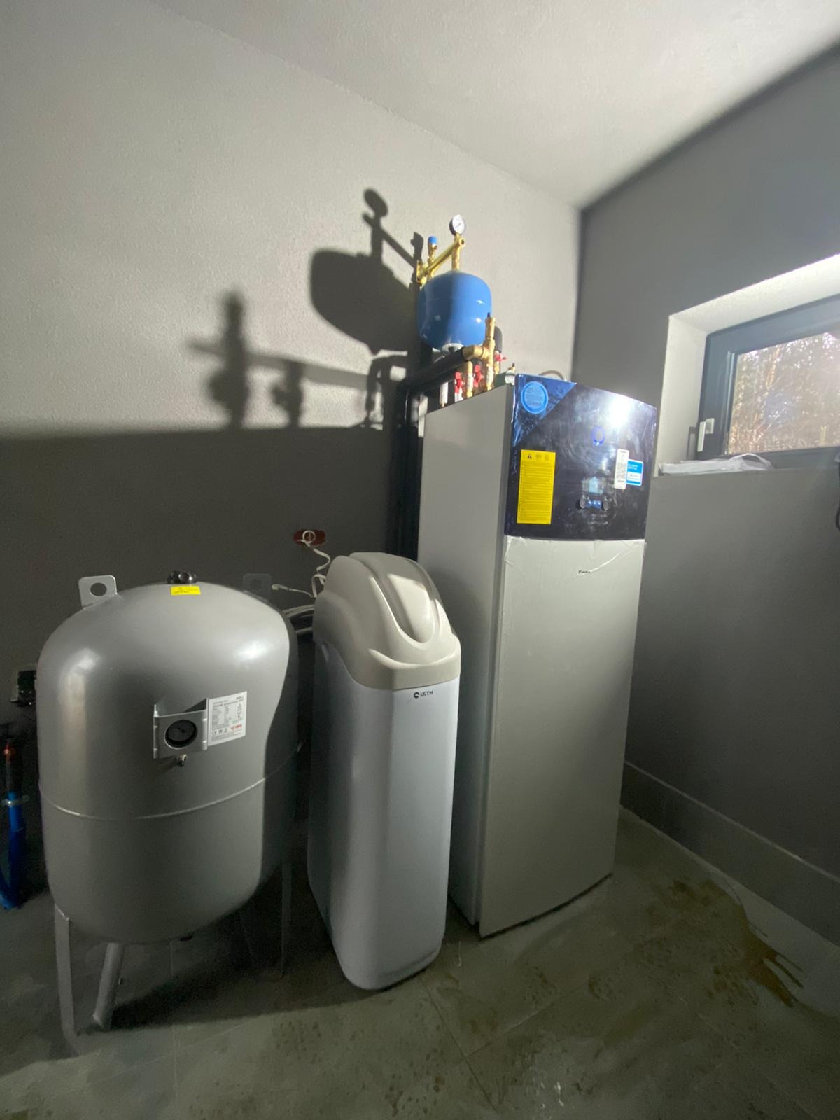 Zestaw urządzeń w kotłowni: pompa ciepła Daikin, zbiornik hydroforowy i zmiękczacz wody, widoczne orurowanie i okno w tle.