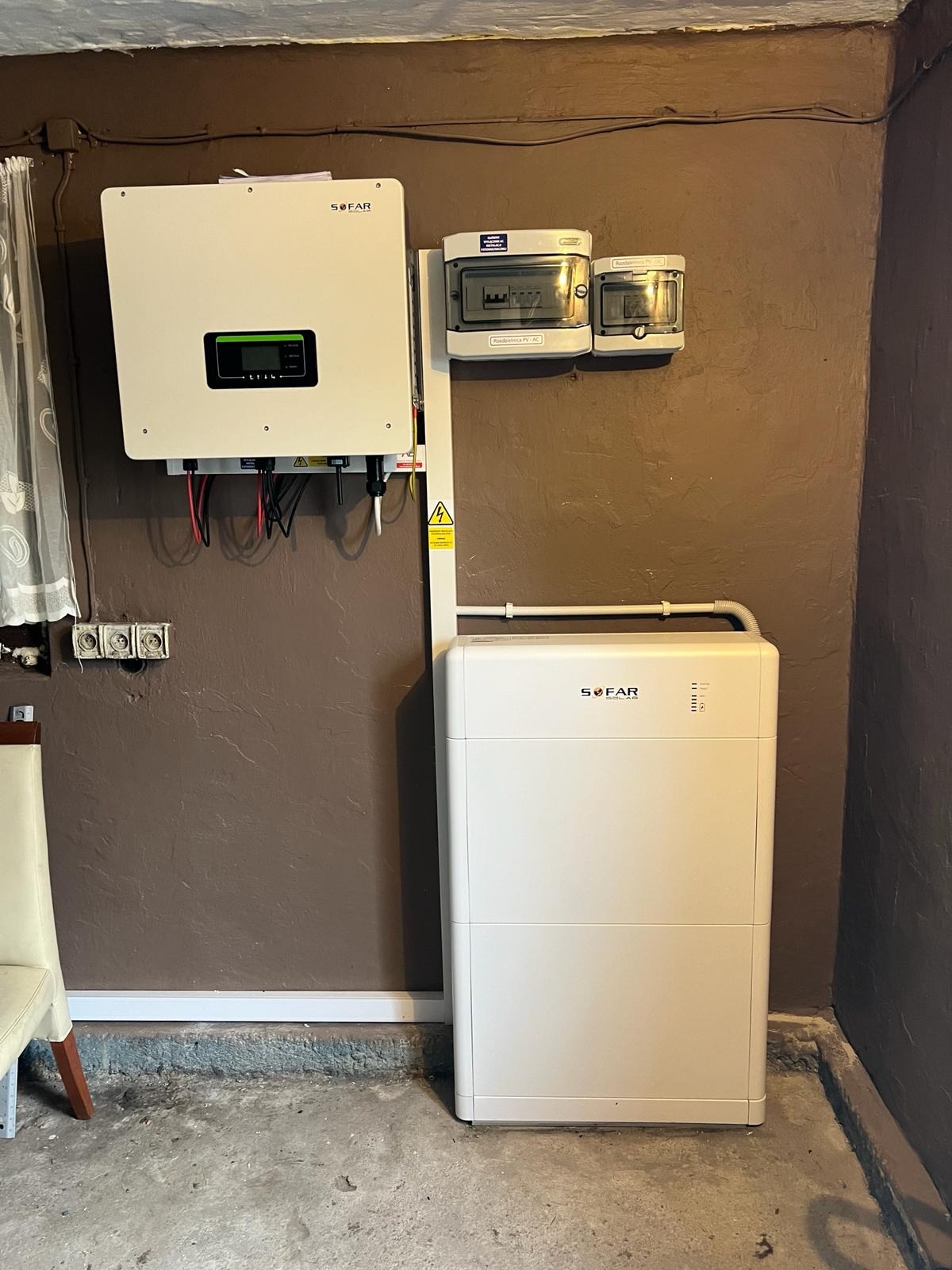 Biały magazyn energii Sofar z podłączonym inwerterem i zabezpieczeniami, zamontowany na brązowej ścianie w piwnicy, widoczne okablowanie i elementy instalacji elektrycznej.