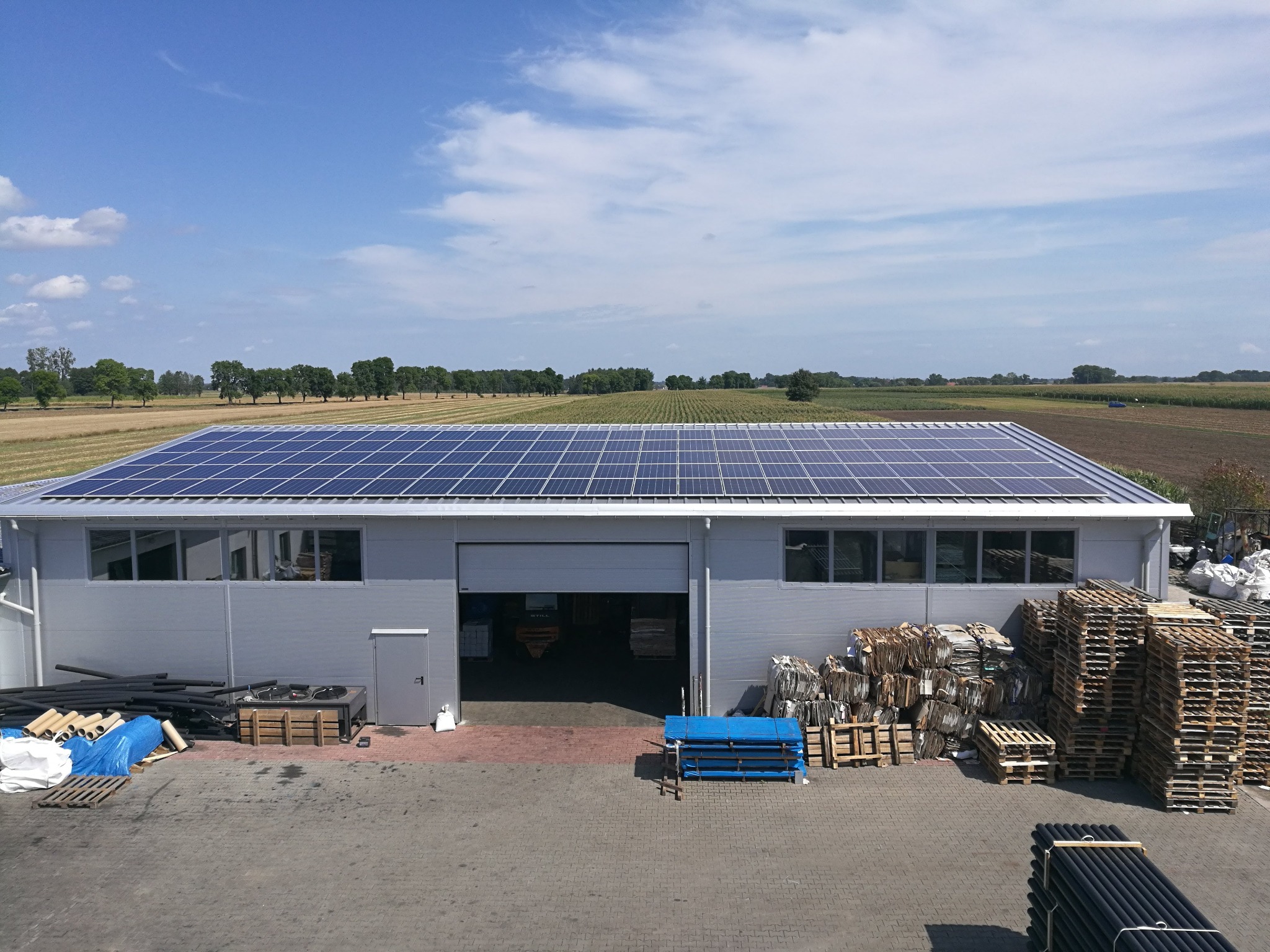 Hala magazynowa z panelami fotowoltaicznymi na dachu, widok z góry na zaplecze z paletami i materiałami budowlanymi, pola uprawne w tle.