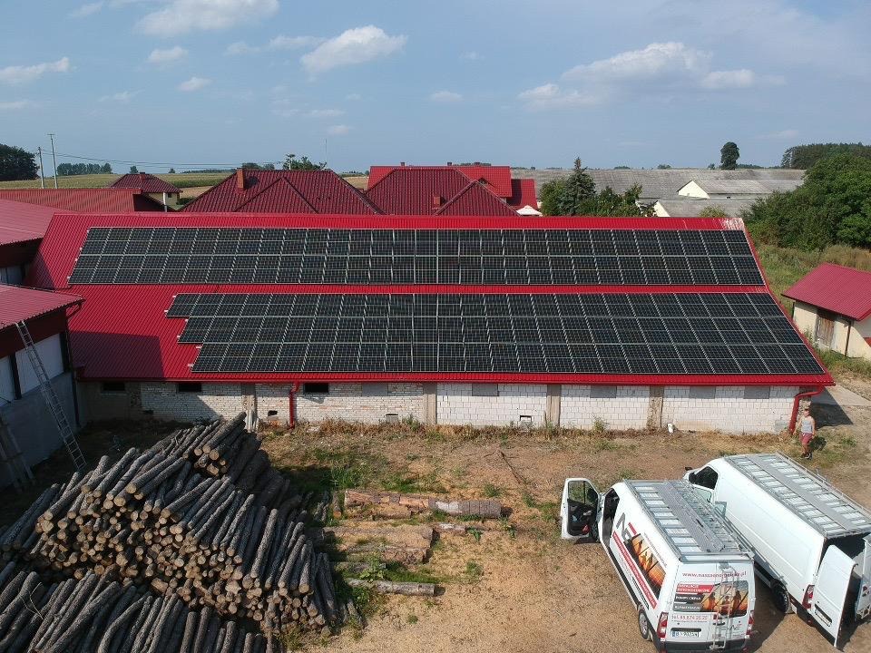 Instalacja paneli fotowoltaicznych na czerwonym dachu budynku gospodarczego, widoczne dwa białe busy z otwartymi drzwiami, sterta pni drzew i osoba w tle.