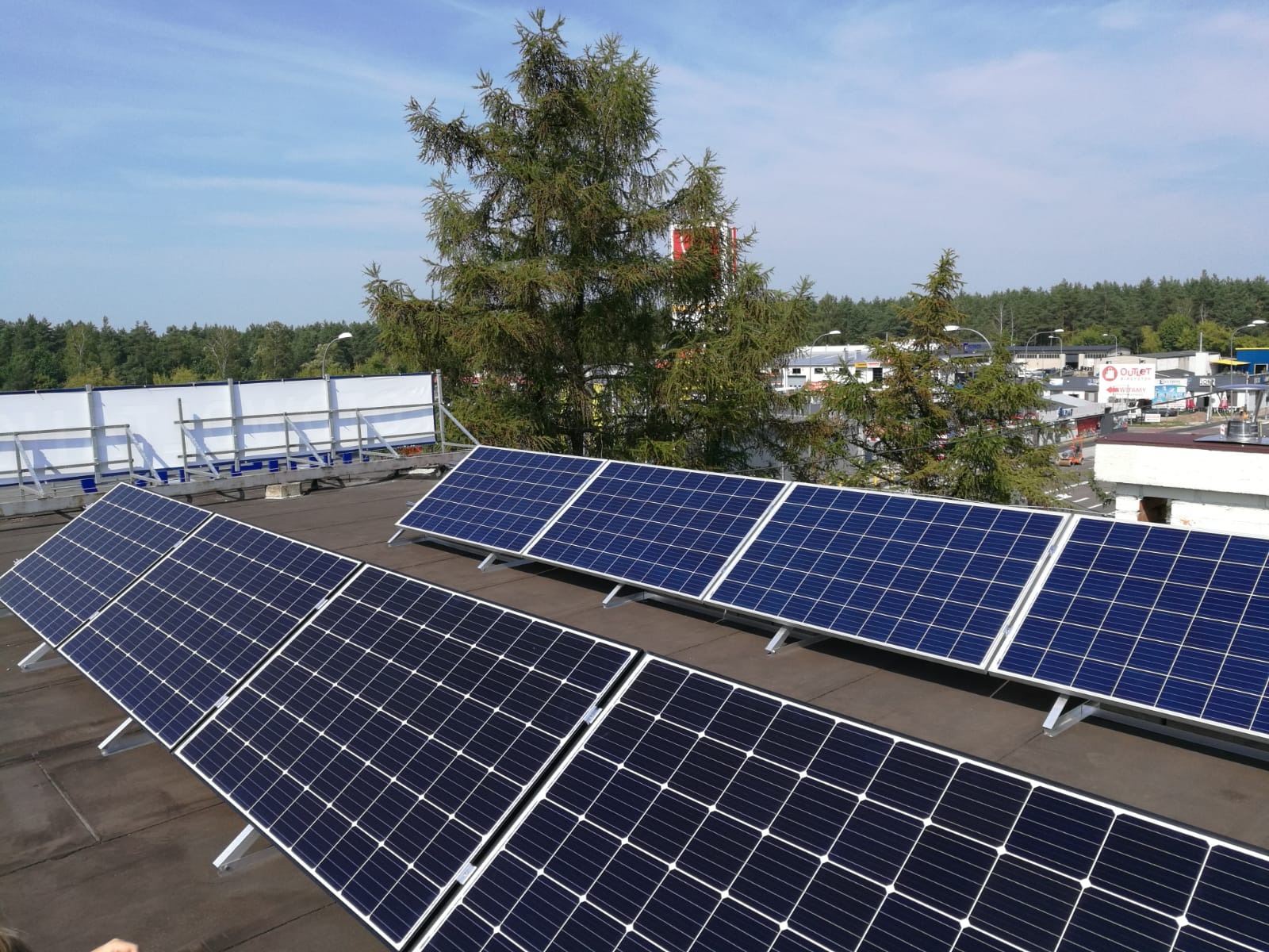 Panel słoneczny na dachu budynku, widok z góry, w tle drzewa i zabudowania miejskie.