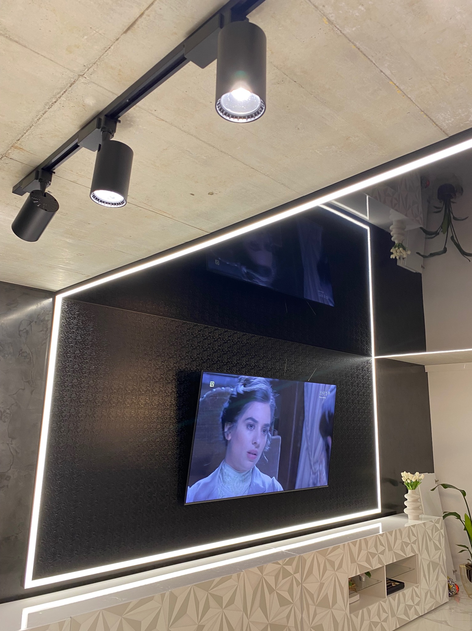 Nowoczesny salon z czarną ścianą, oświetleniem LED wzdłuż krawędzi i reflektorami na szynie podsufitowej, z telewizorem wyświetlającym film kostiumowy.