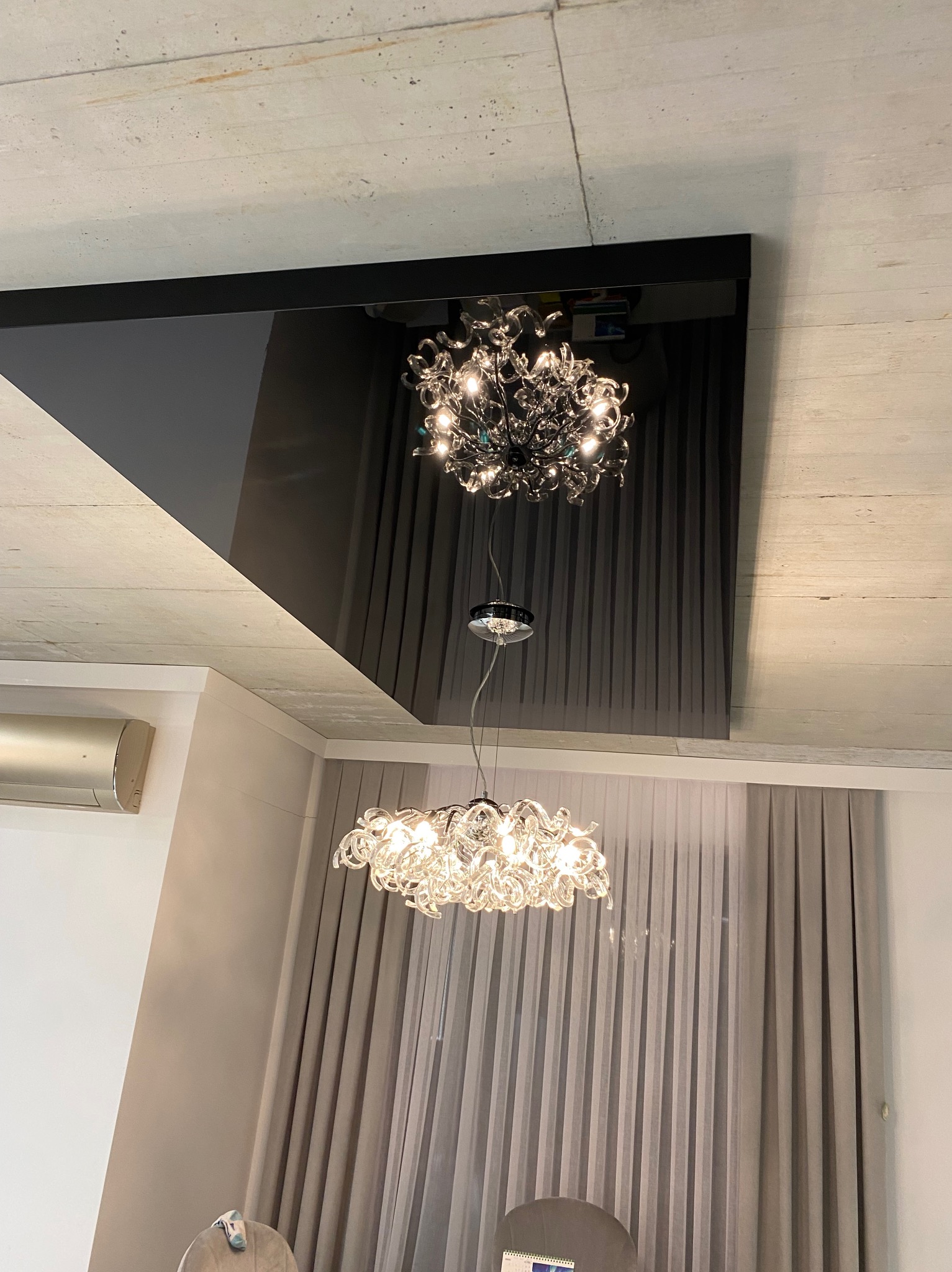 Nowoczesny salon z dwoma żyrandolami: jeden odbija się w błyszczącym, czarnym panelu sufitowym, drugi zwisa poniżej, oświetlając szare zasłony i część pomieszczenia z klimatyzatorem.