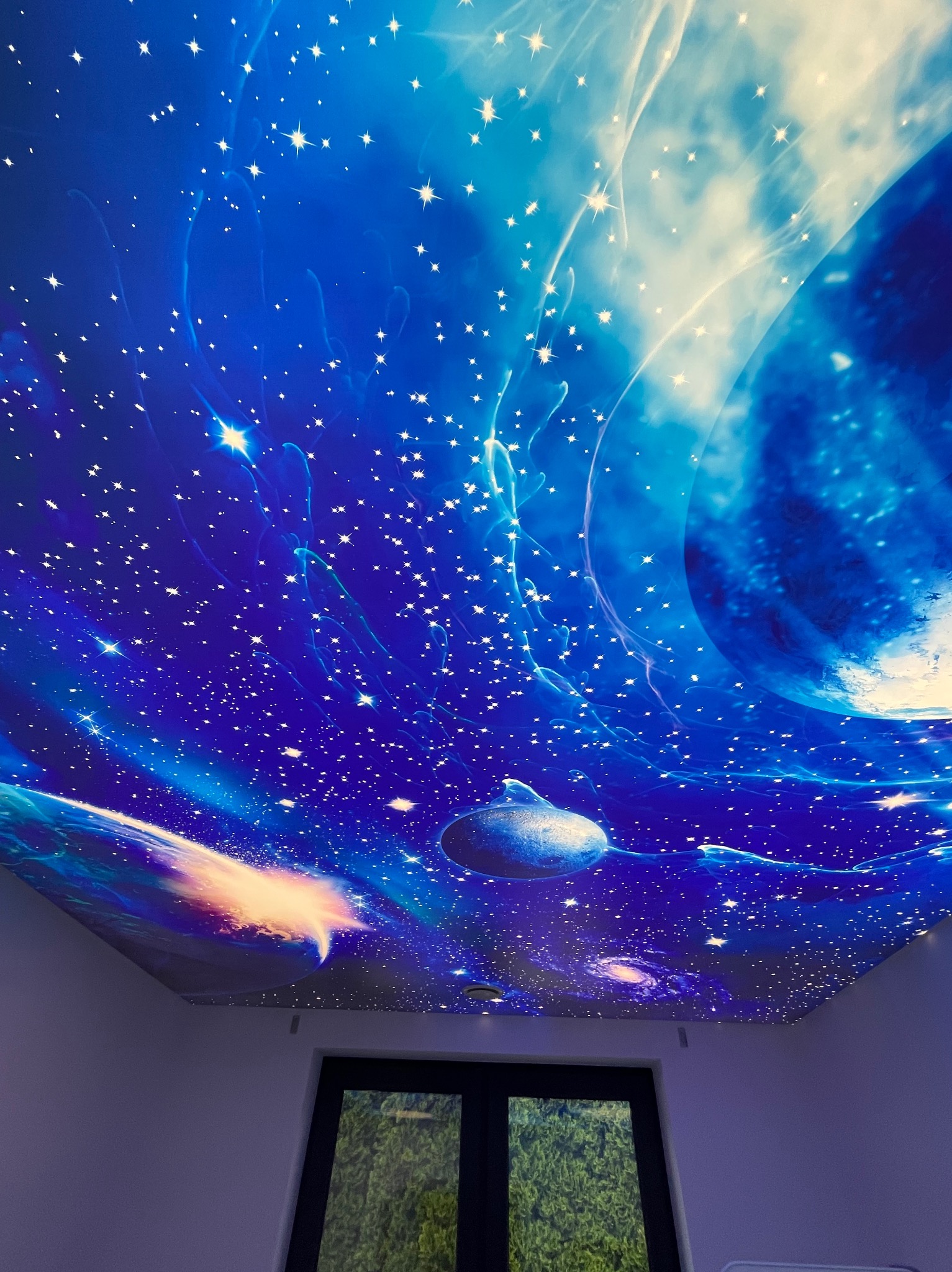 Niebieski sufit z motywem kosmosu i gwiazd, widok z dołu, z oknem w tle. Dekoracyjny motyw galaktyki, oświetlenie LED.