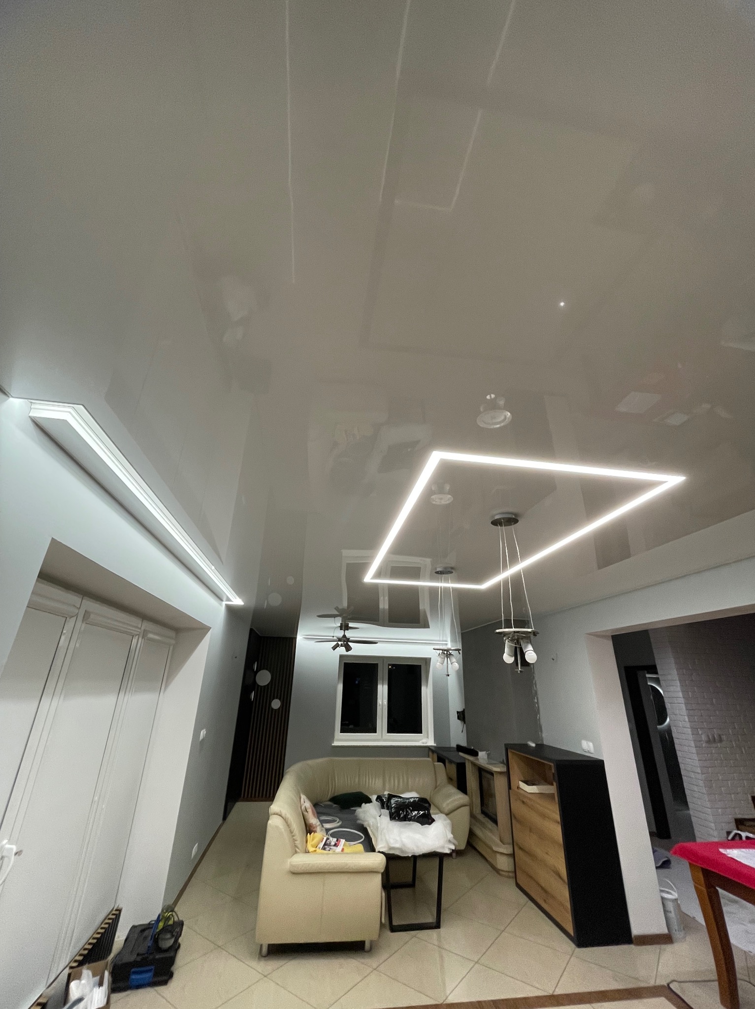 Błyszczący, biały sufit z oświetleniem LED w kształcie kwadratu, wiszące lampy, beżowa kanapa. Widok na pokój z perspektywy sufitu.