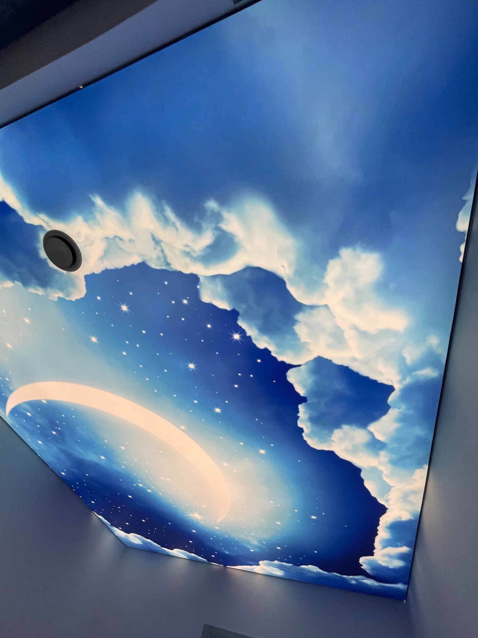 Nastrojowy sufit z motywem nieba: błękit, chmury i gwiazdy. Okrągły otwór wentylacyjny wkomponowany w design, tworzy unikalny efekt wizualny w pomieszczeniu.