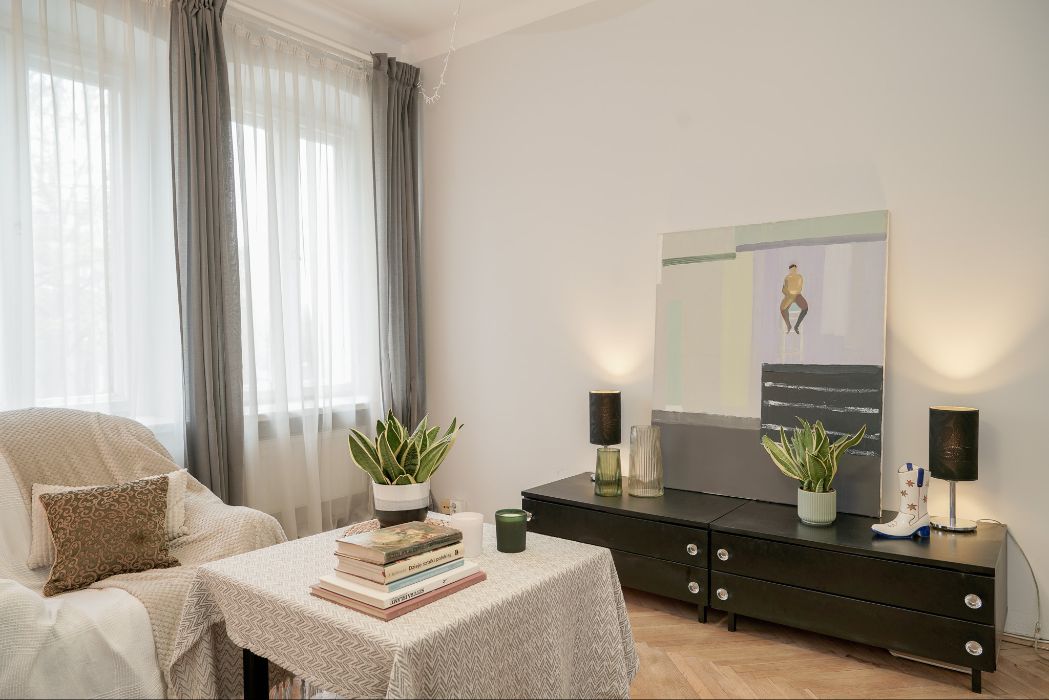 Jasne wnętrze mieszkania z oknem, zasłonami, obrazem, stolikiem z książkami i roślinami. Parkiet na podłodze. Lampki nocne na komodzie.
