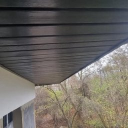 PokryjemyCiDach PATRYK BOBIŃSKI - Podbitka dachowa z ciemnego drewna zamontowana pod okapem budynku z jasną elewacją, widok na drzewa w tle.