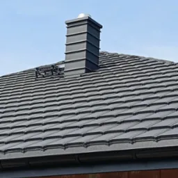 Nowy dach z ciemnoszarej dachówki ceramicznej z kominem systemowym w kolorze, widoczny fragment rynny i bariera przeciwśniegowa.