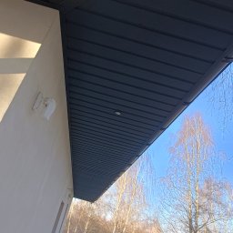 PokryjemyCiDach PATRYK BOBIŃSKI - Nowoczesne podbitka dachowa w kolorze grafitowym z wbudowanym oświetleniem punktowym, widok z dołu na tle błękitnego nieba i drzew bez liści.