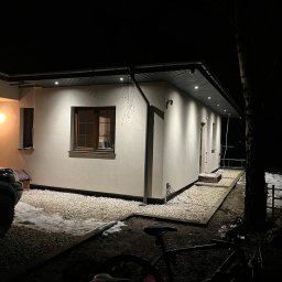 PokryjemyCiDach PATRYK BOBIŃSKI - Elewacja domu jednorodzinnego oświetlona nocą, widoczne rynny i obróbki blacharskie, teren wokół wysypany żwirem, częściowo pokryty śniegiem, z elementami roweru na pierwszym planie.