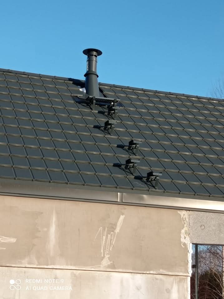 Szary dach z dachówki ceramicznej z zamontowanymi stopniami kominiarskimi i kominem w kolorze antracytowym na tle błękitnego nieba.