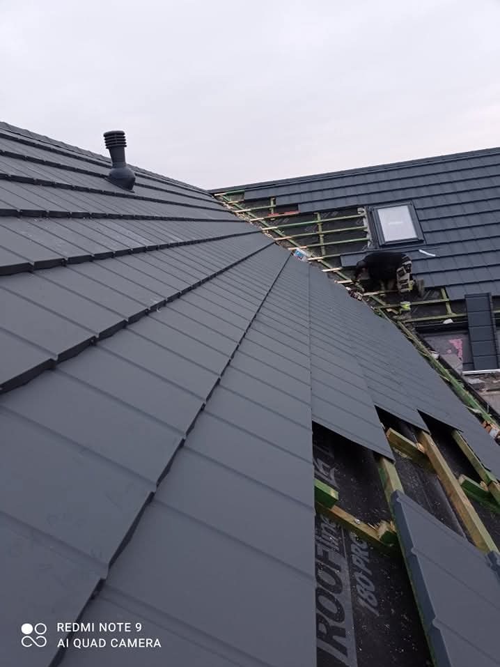 Montaż szarej blachodachówki na dachu domu jednorodzinnego, widoczny kominek wentylacyjny, okno dachowe oraz fragment konstrukcji dachu z membraną w trakcie prac.
