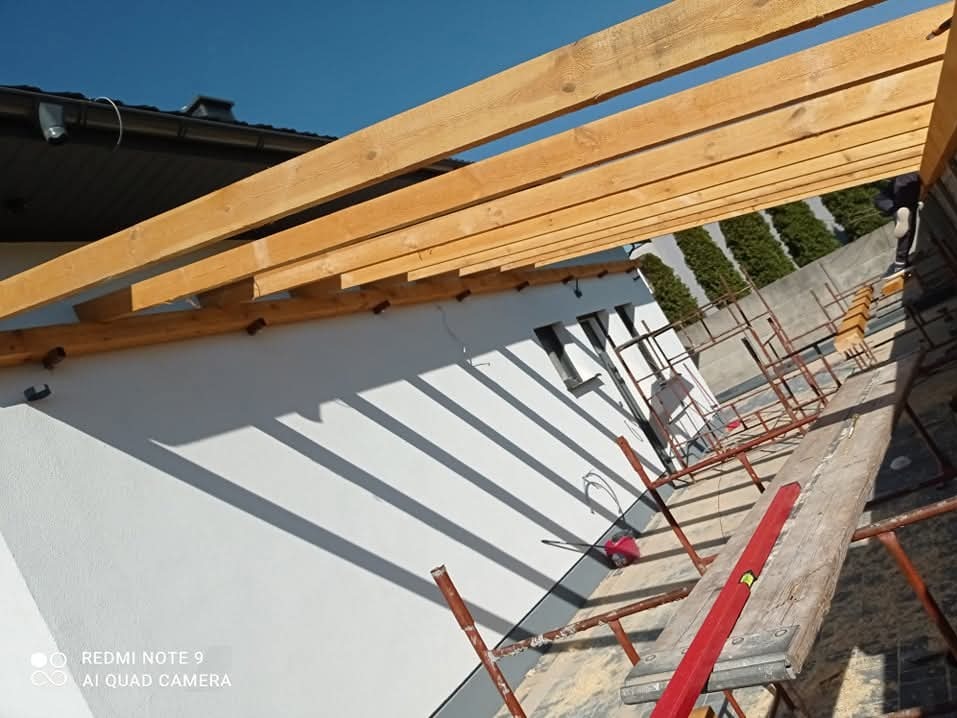Montaż drewnianej więźby dachowej na białym budynku, widoczne rusztowanie i poziomica, błękitne niebo w tle.