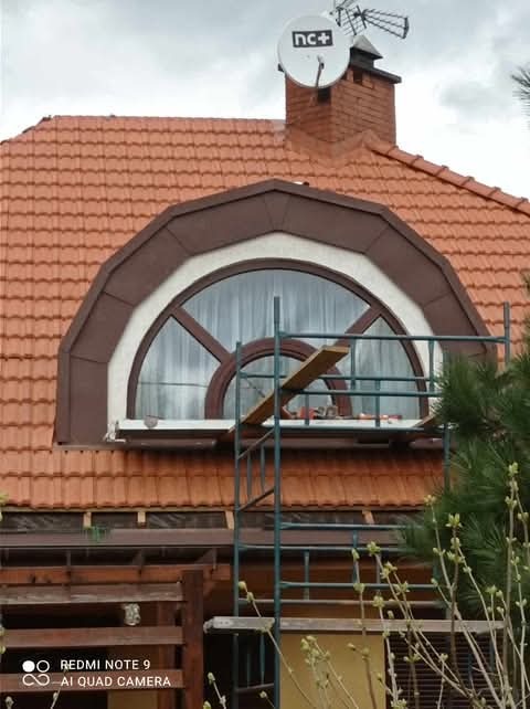 Dach pokryty czerwoną dachówką ceramiczną z oknem mansardowym w brązowej ramie, na dachu widoczna antena satelitarna, rusztowanie przed oknem.