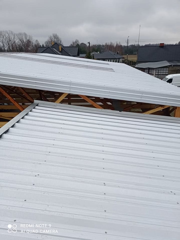 Konstrukcja dachu z drewnianą więźbą i jasnym, metalowym pokryciem w trakcie budowy; w tle widoczne inne budynki i pochmurne niebo.