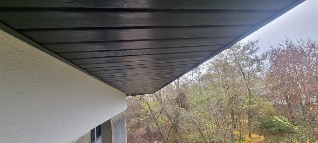 Podbitka dachowa z ciemnego drewna zamontowana pod okapem budynku z jasną elewacją, widok na drzewa w tle.