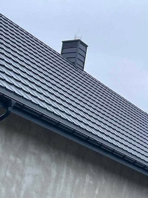 Nowy, grafitowy dach z metalowej dachówki modułowej, z widocznym kominem w tym samym kolorze i systemem rynnowym, zamontowany na budynku o szarej elewacji.