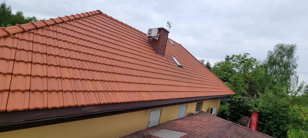 Czerwona dachówka ceramiczna na skośnym dachu domu z kominem, na którym zamontowano klimatyzator. Widoczna rynna w kolorze ciemnego brązu, okna dachowe i fragment elewacji budynku w kolorze żółtym,...