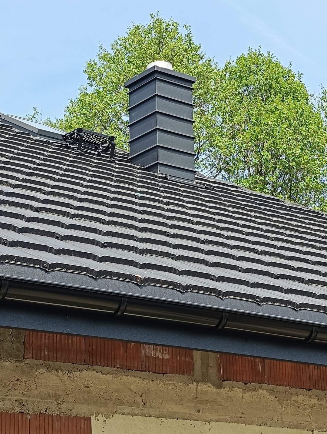 Ciemnoszara dachówka ceramiczna na nowym dachu z kominem z blachy trapezowej i zamontowanymi rynnami, widok z dołu na tle zielonych drzew.