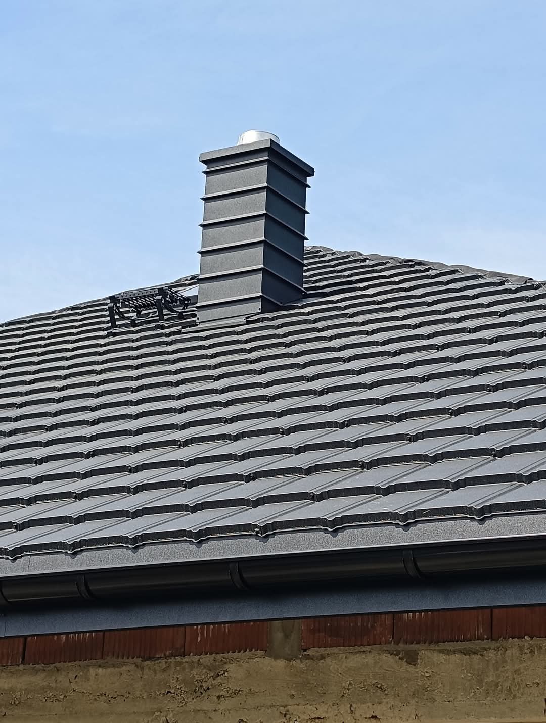 Nowy dach z ciemnoszarej dachówki ceramicznej z kominem systemowym w kolorze, widoczny fragment rynny i bariera przeciwśniegowa.