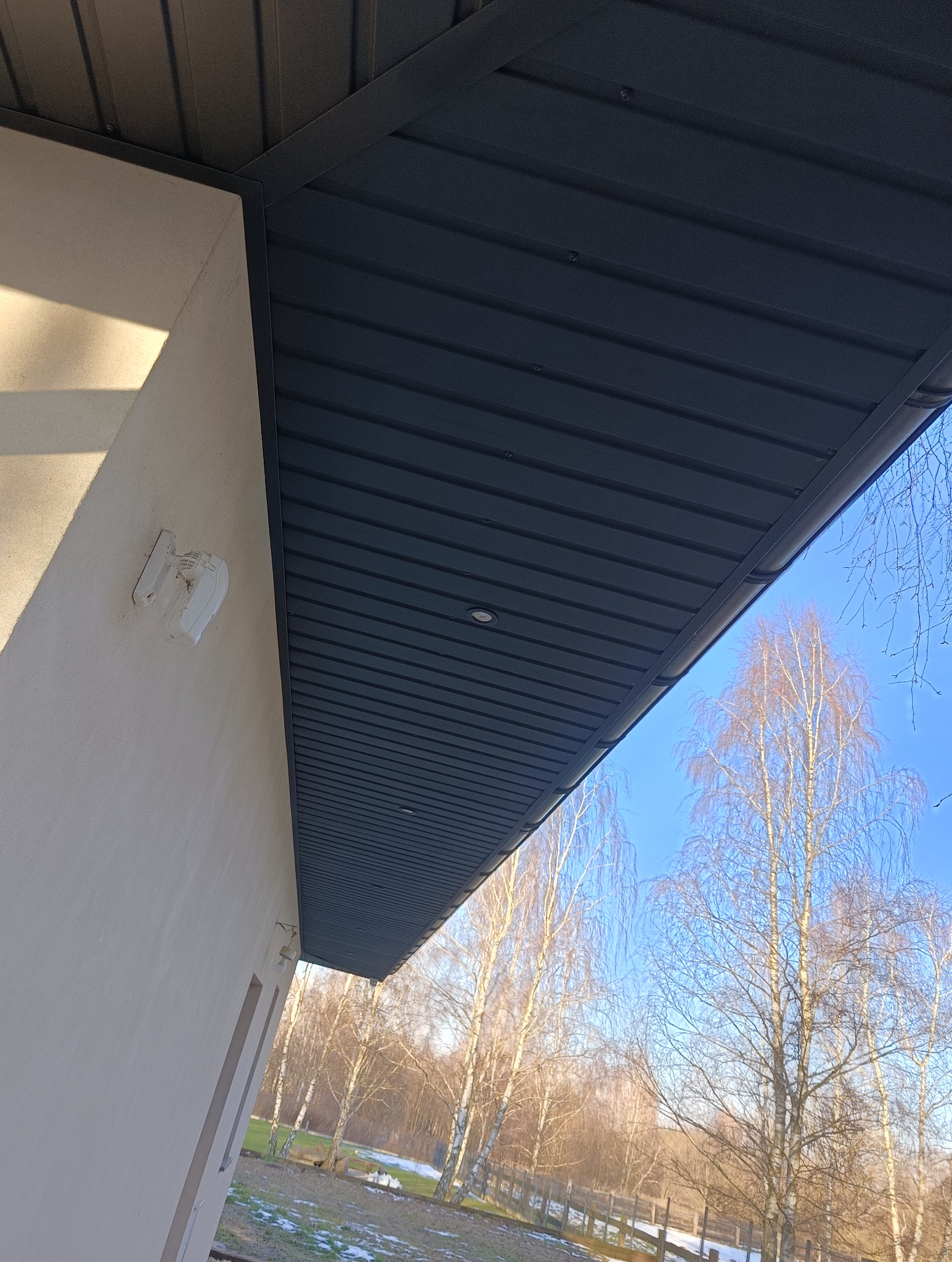 Nowoczesne podbitka dachowa w kolorze grafitowym z wbudowanym oświetleniem punktowym, widok z dołu na tle błękitnego nieba i drzew bez liści.