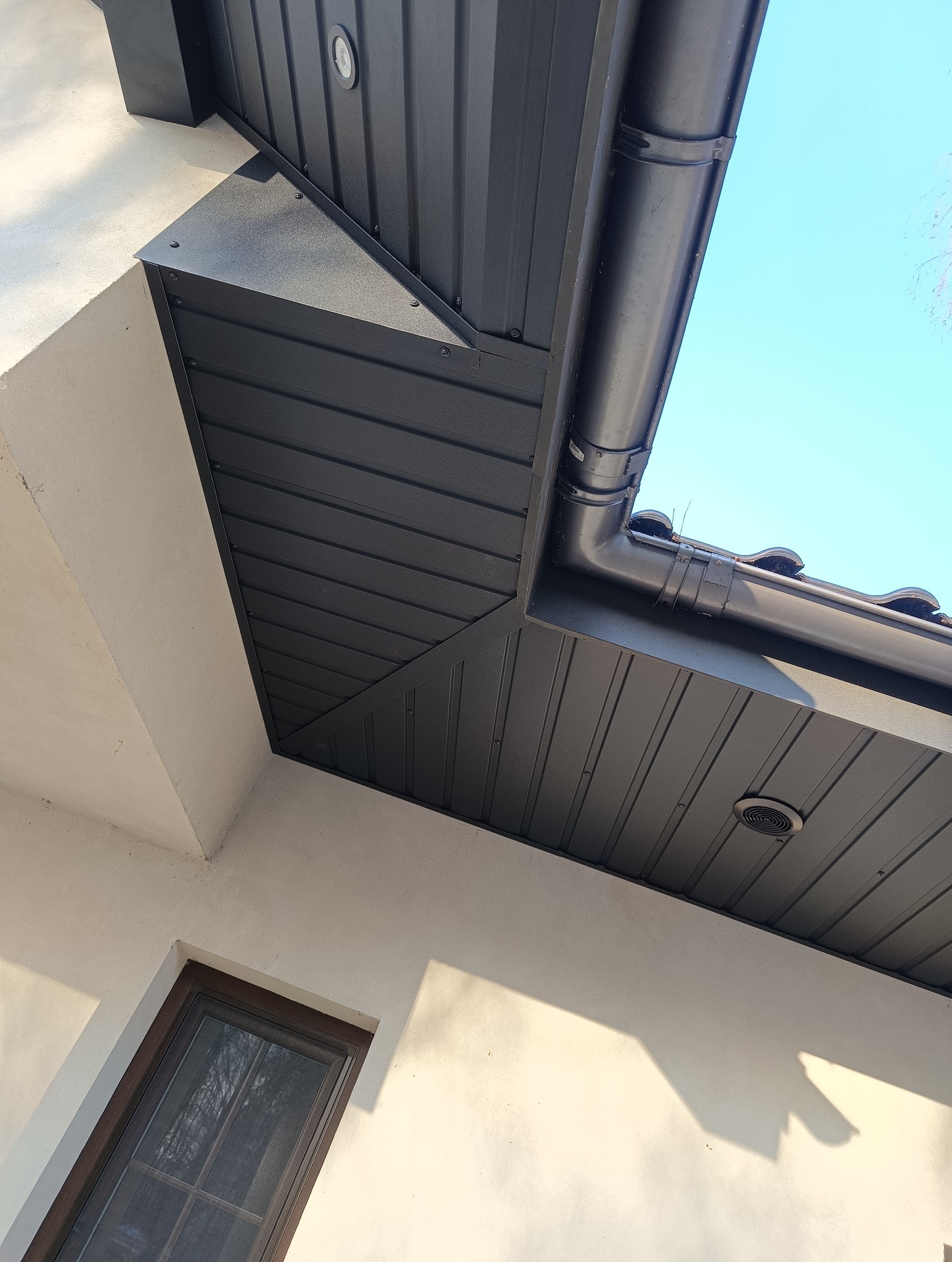 Nowoczesny, czarny system orynnowania zamontowany na podbitce dachu, widok z dołu na tle błękitnego nieba.