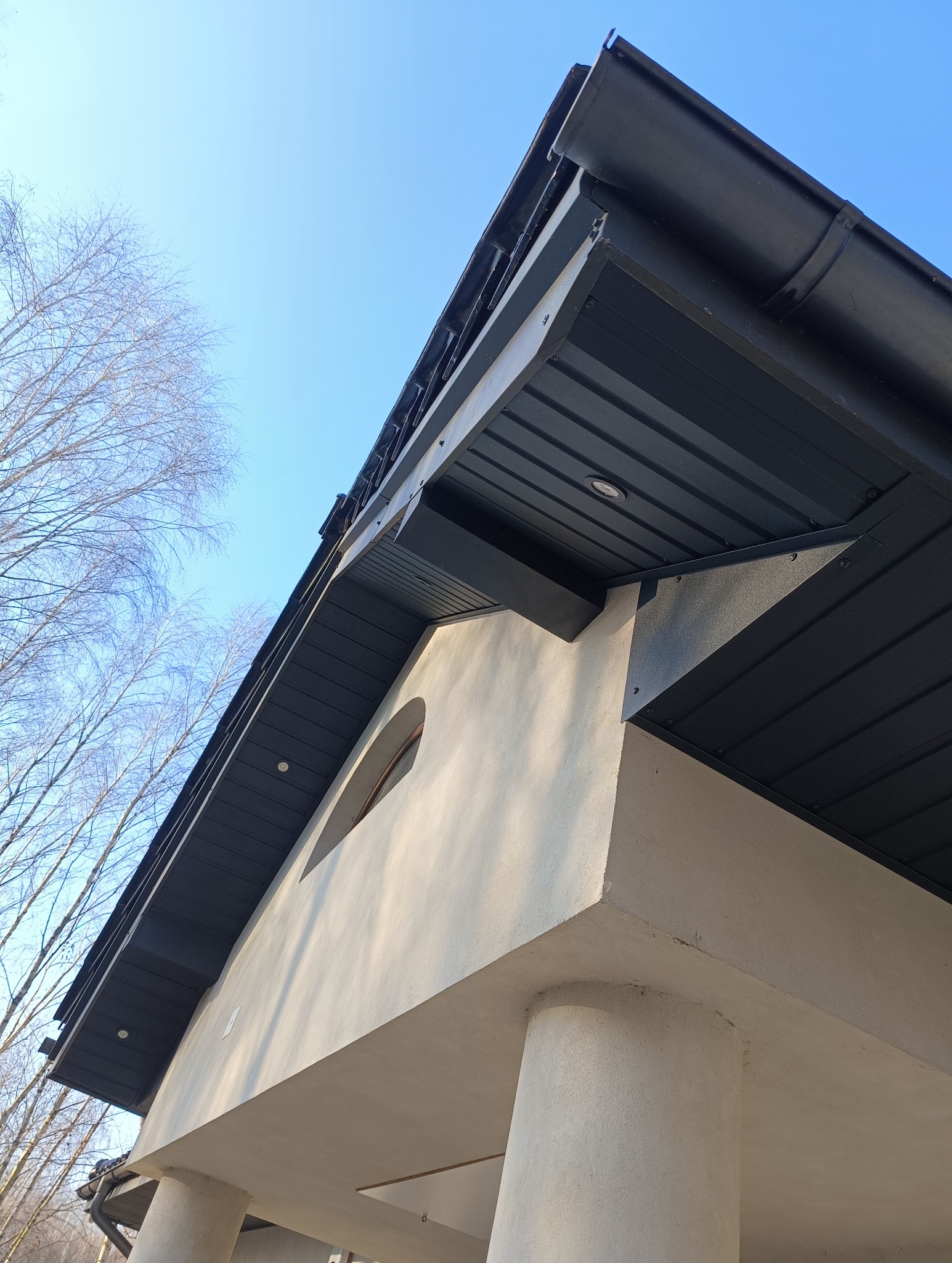Fragment dachu z blachy trapezowej w kolorze grafitowym, widok z dołu na okap z oświetleniem punktowym, fragment elewacji z okrągłym oknem i kolumny podtrzymujące zadaszenie, błękitne niebo...