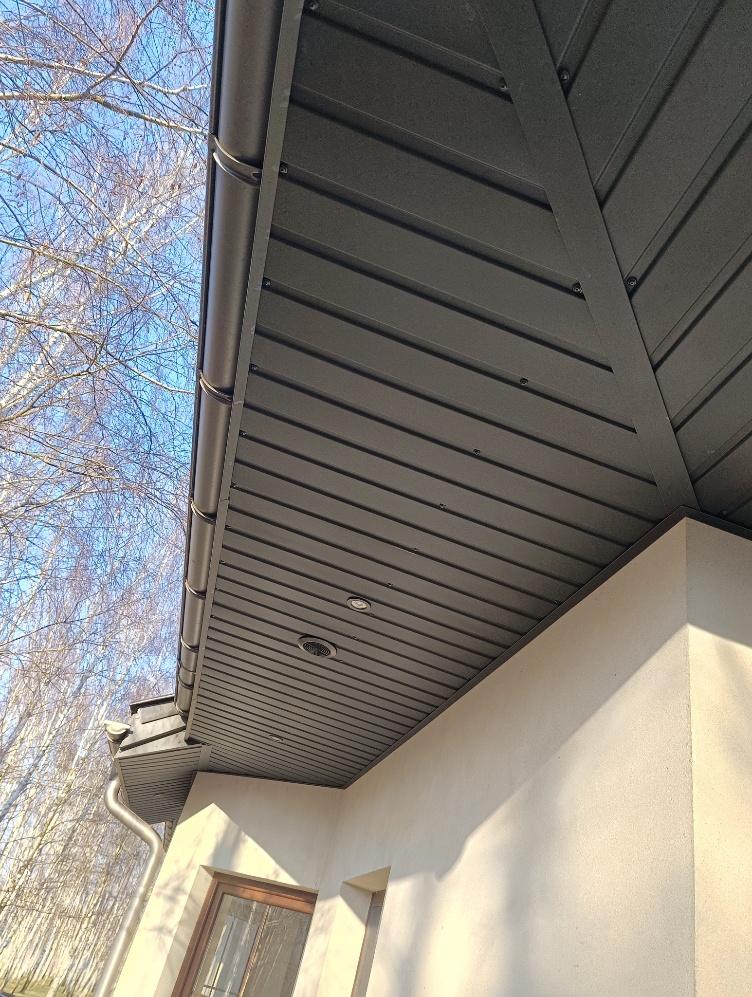 Nowoczesny dach z brązowej blachy trapezowej, widok z dołu na podbitkę z oświetleniem punktowym i rynnę, na tle błękitnego nieba z gałęziami drzew.