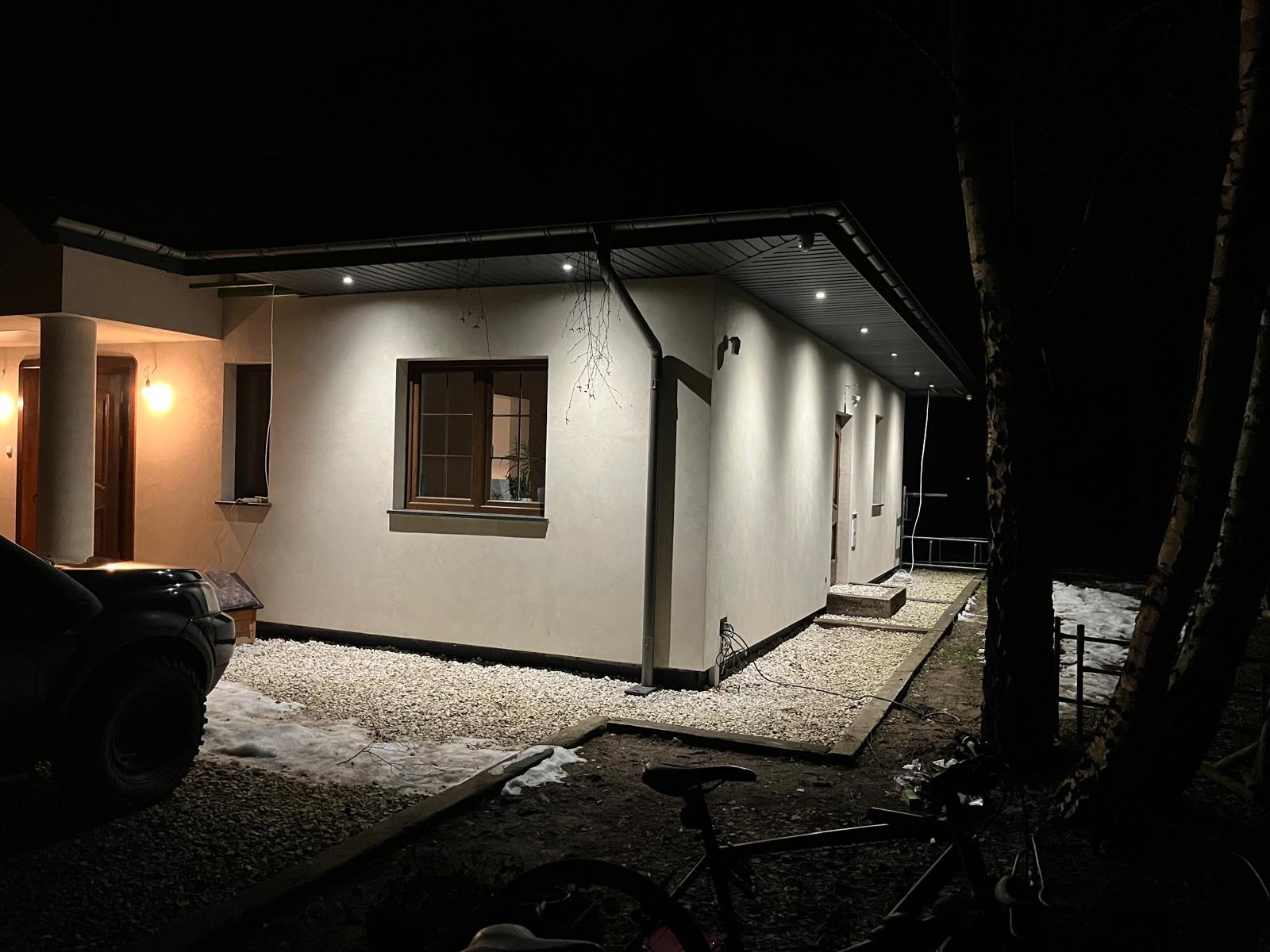Elewacja domu jednorodzinnego oświetlona nocą, widoczne rynny i obróbki blacharskie, teren wokół wysypany żwirem, częściowo pokryty śniegiem, z elementami roweru na pierwszym planie.