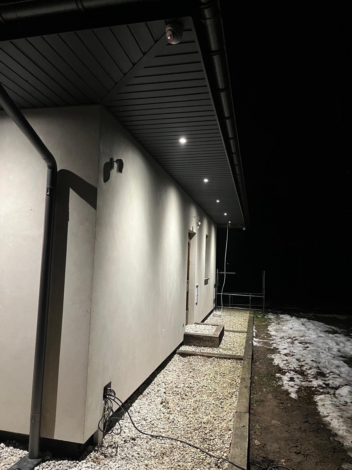 Elewacja budynku oświetlona punktowymi lampami pod okapem, widoczny system rynnowy i kamera monitoringu, nocne ujęcie.