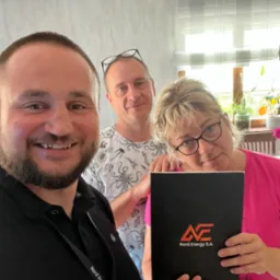 Trzy osoby pozują do zdjęcia w pomieszczeniu, jedna z nich trzyma czarny folder z logo Nord Energy S.A.