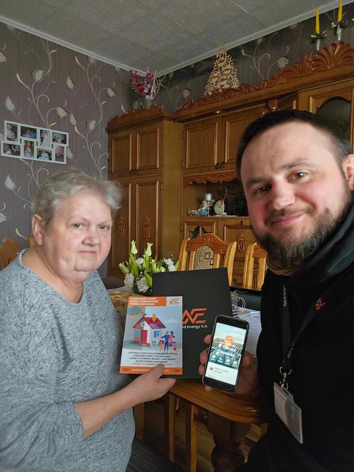 Kobieta i mężczyzna prezentują materiały reklamowe firmy Nord Energy S.A. dotyczące oszczędności energii w domu, w tle widoczny jest klasyczny wystrój wnętrza.