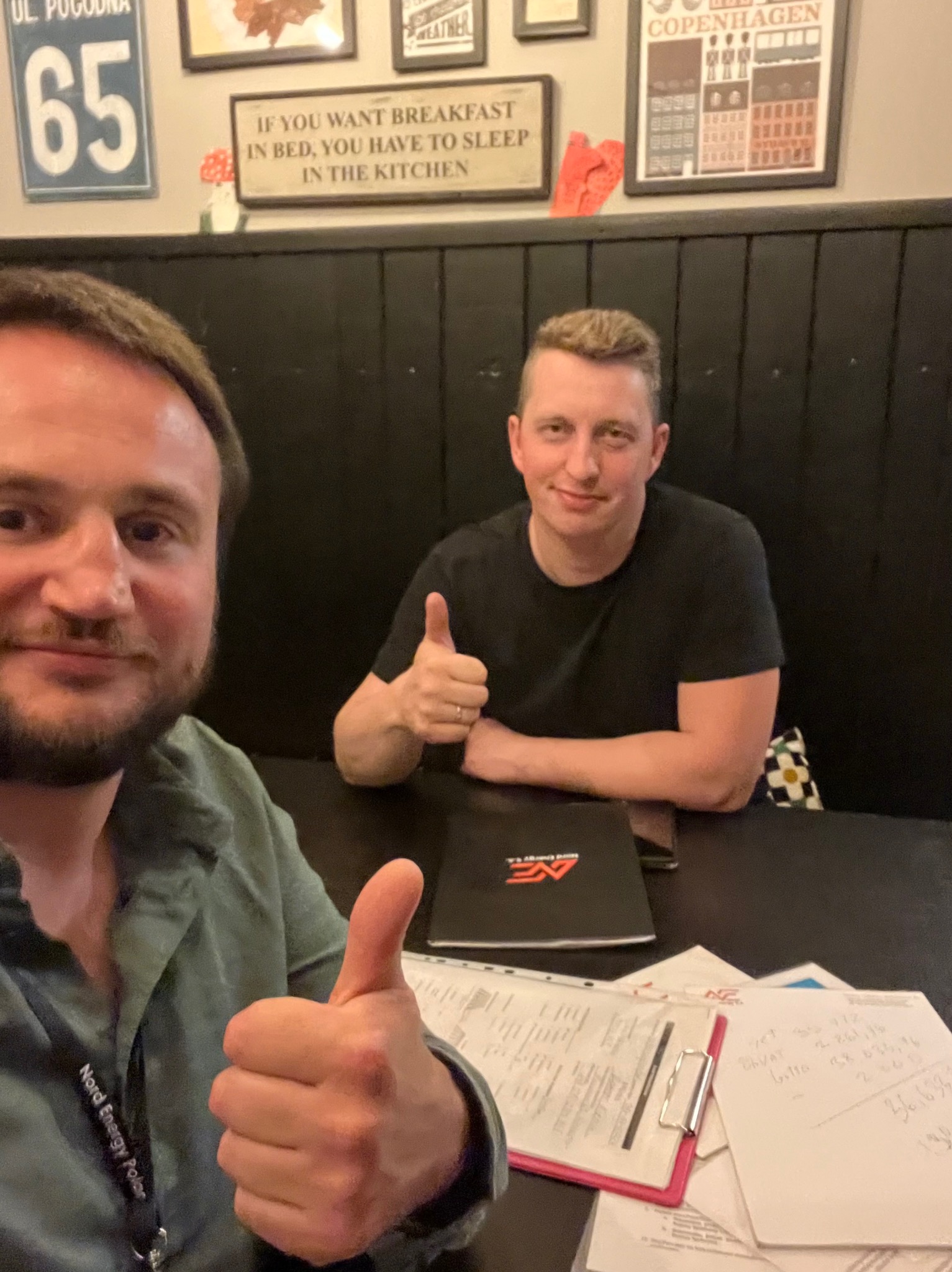 Dwóch mężczyzn siedzi przy stole w restauracji, obaj pokazują kciuki w górę. Na stole leżą dokumenty, notatki i firmowy folder Nord Energy Polar.