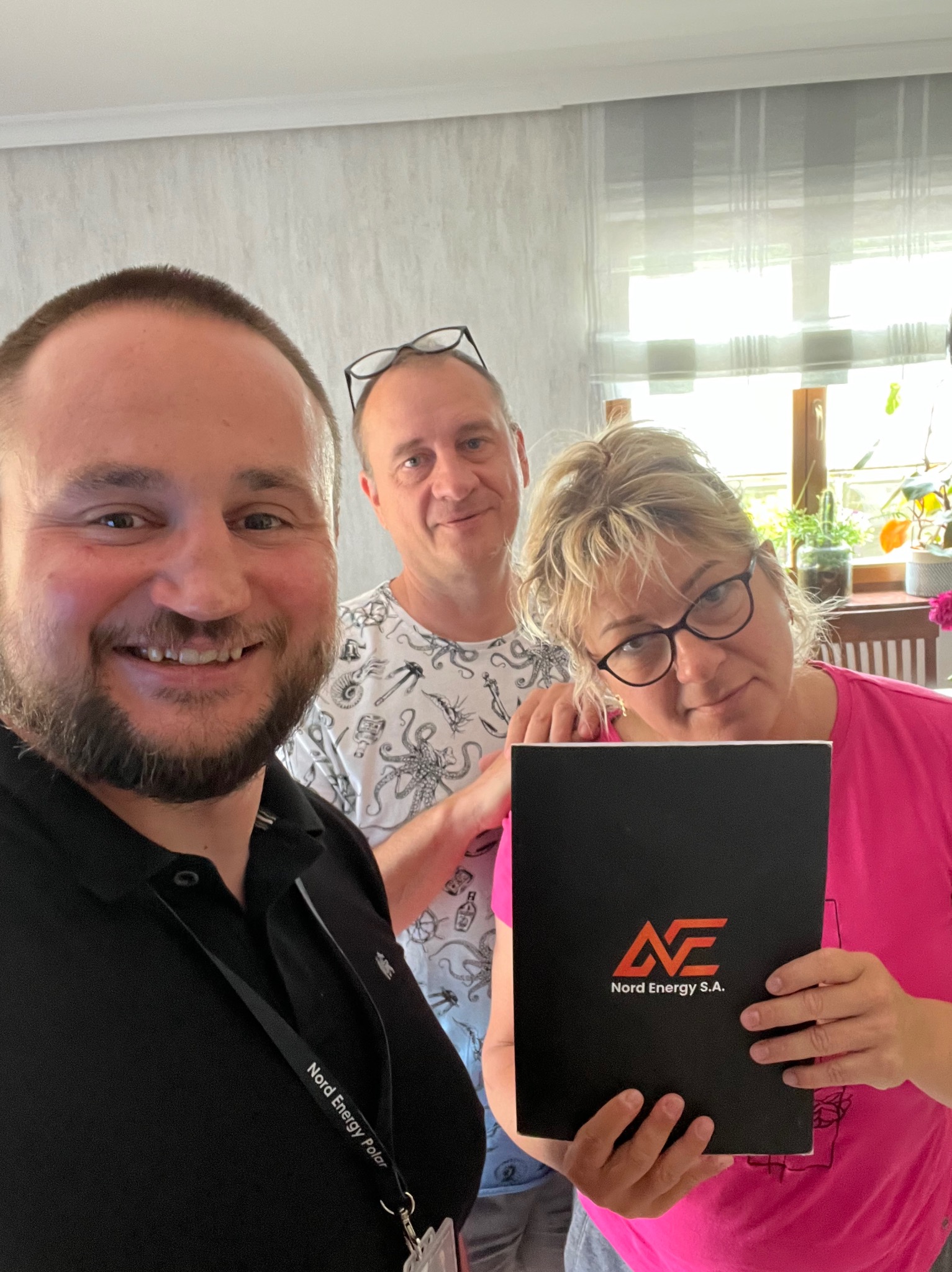 Trzy osoby pozują do zdjęcia w pomieszczeniu, jedna z nich trzyma czarny folder z logo Nord Energy S.A.