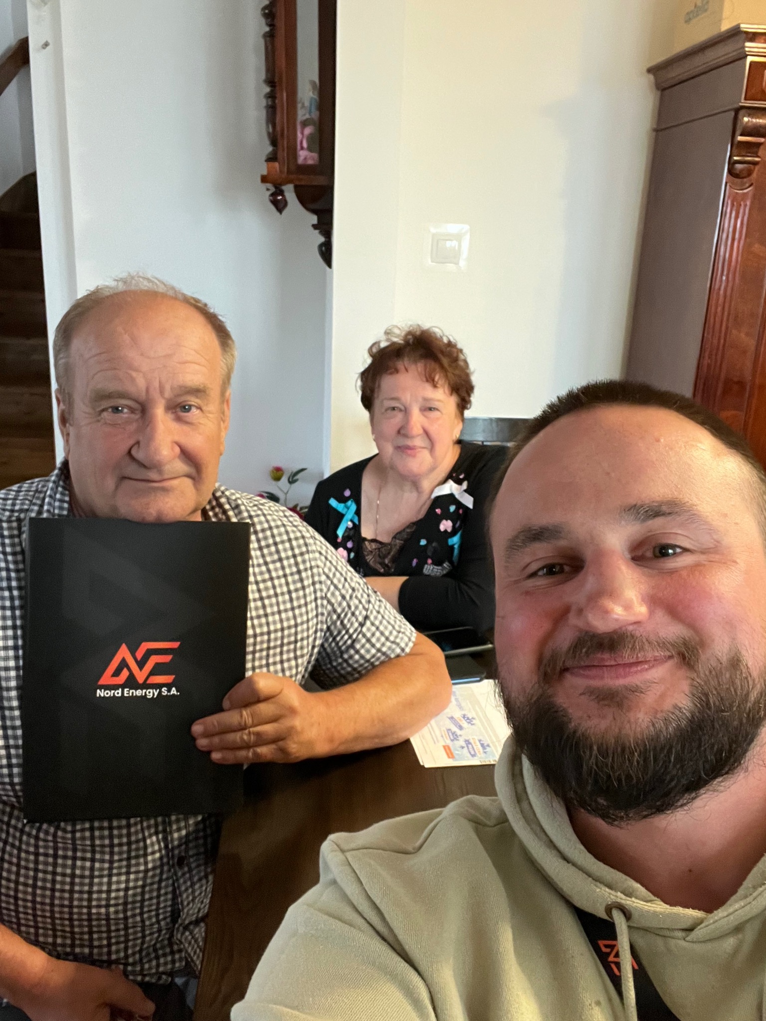 Trzy osoby pozują do selfie przy stole, jedna trzyma czarny folder z logo Nord Energy S.A.